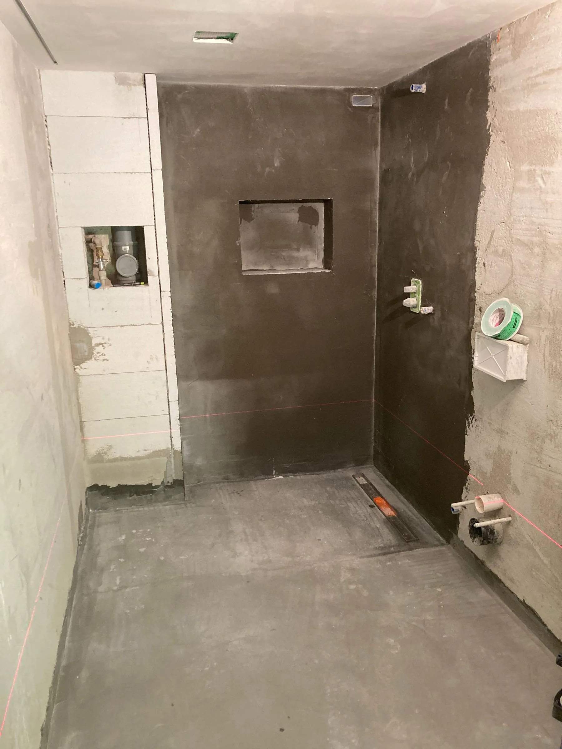 Sprcha před obložením: tmavá hydroizolace na podlaze i stěnách, žlab je osazený