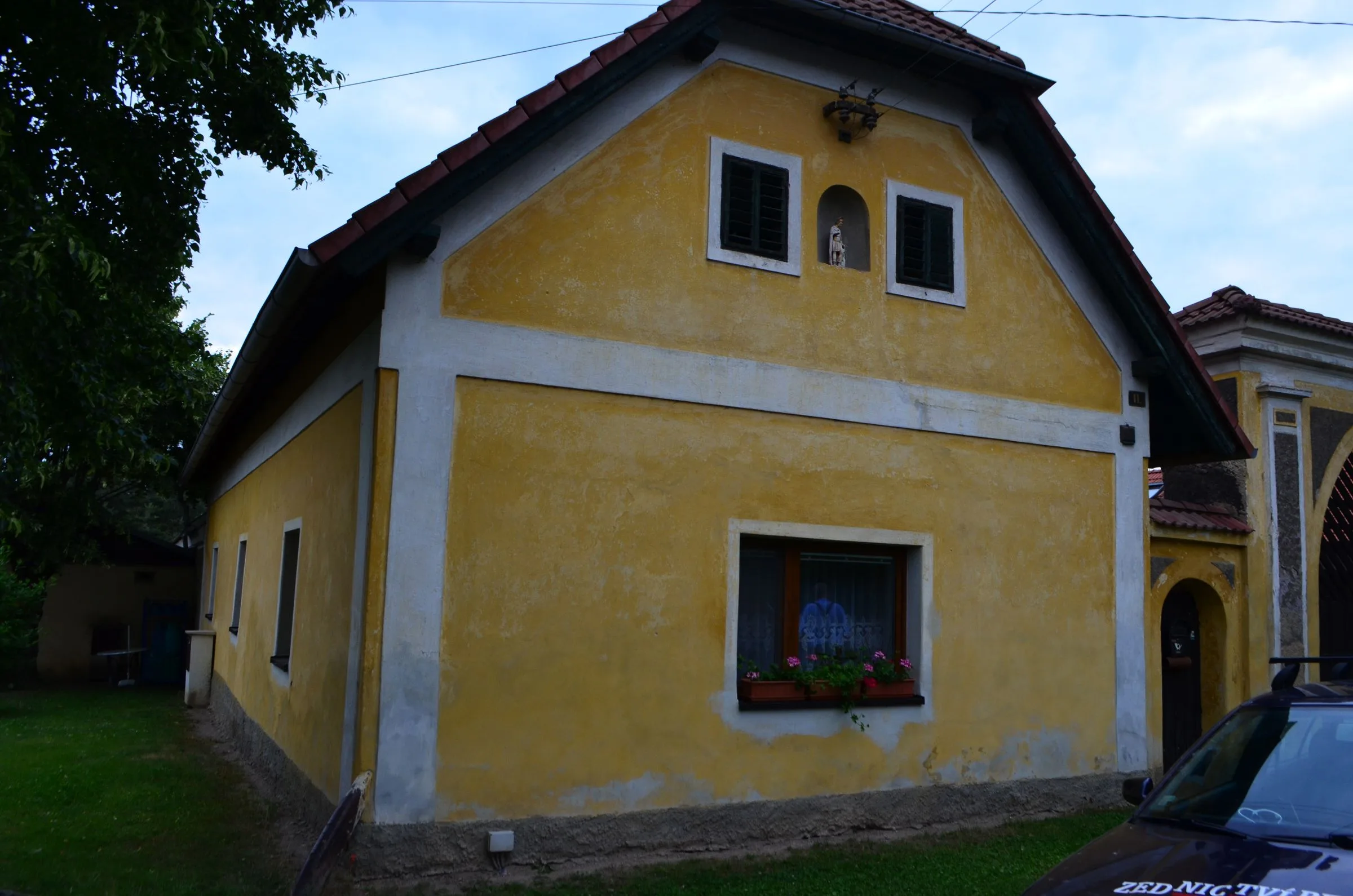 Štít domu se starou oprýskanou žlutou omítkou a&nbsp;nikou se soškou